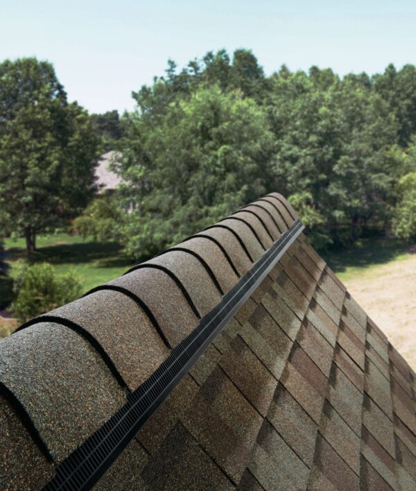 A close-up view of a house roof peak with brown shingles and a black ridge vent, with green trees and a lawn visible in the background under a clear sky. - RoofZen, Chester County PA Roofing, Siding and More