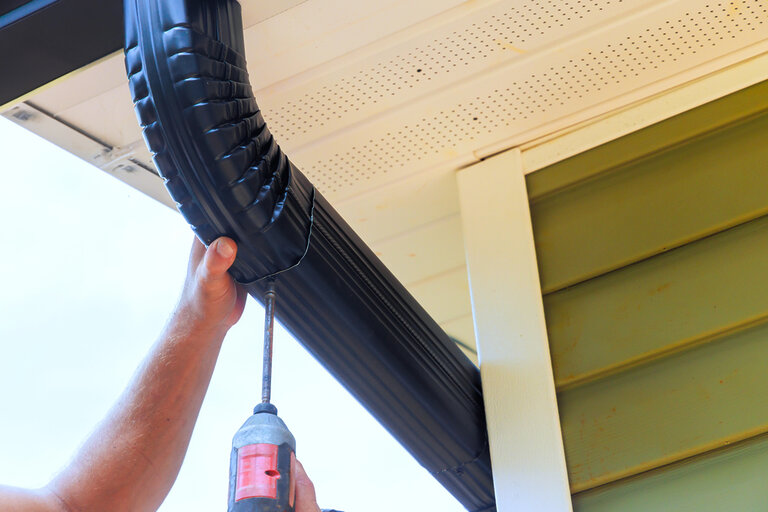 A person uses a power drill to install or secure a black downspout to the gutter system on the corner of a house with green siding and a white soffit. - RoofZen, Chester County PA Roofing, Siding and More