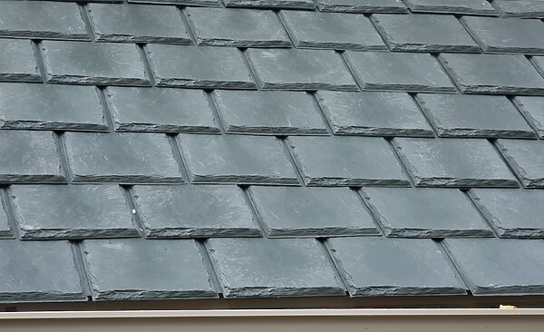 Close-up view of overlapping gray slate roof tiles arranged in neat, horizontal rows with visible texture and edges. A section of the roof edge and gutter is visible at the bottom. - RoofZen, Chester County PA Roofing, Siding and More