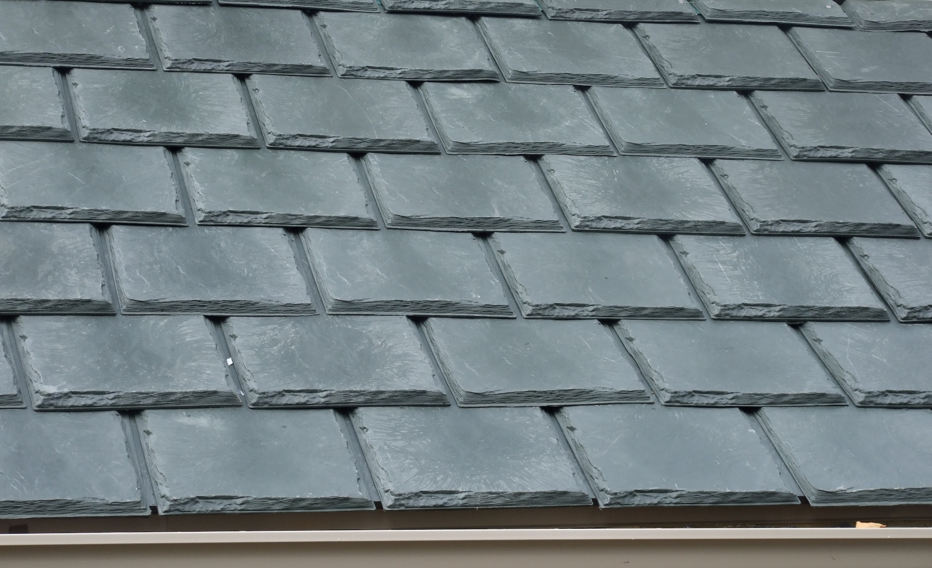 Close-up view of overlapping gray slate roof tiles arranged in neat, horizontal rows with visible texture and edges. A section of the roof edge and gutter is visible at the bottom. - RoofZen, Chester County PA Roofing, Siding and More