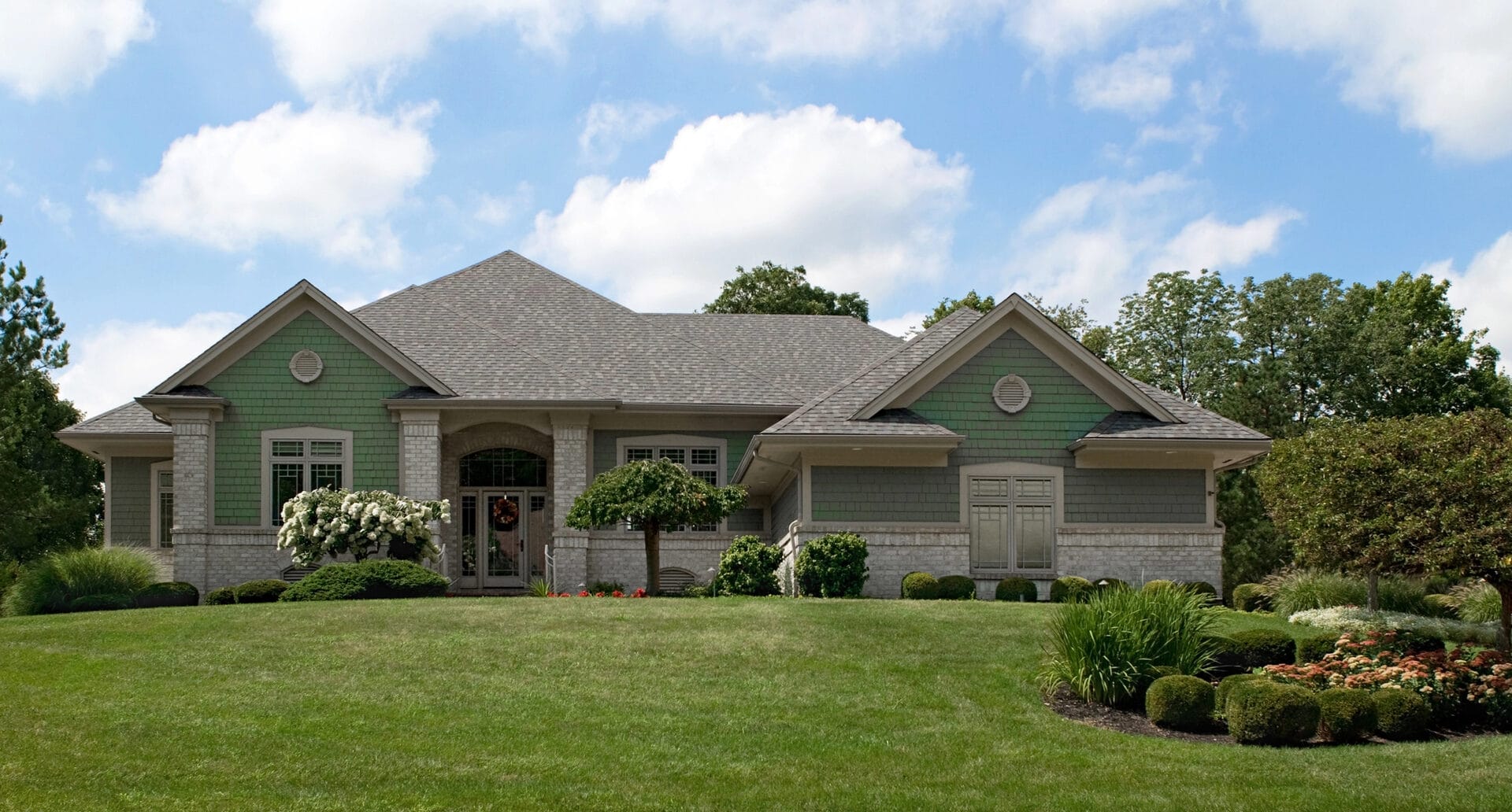 A single-story suburban house with green and beige exterior, arched doorway, large windows, and a well-manicured lawn with shrubs and flowering bushes under a partly cloudy sky. - RoofZen, Chester County PA Roofing, Siding and More