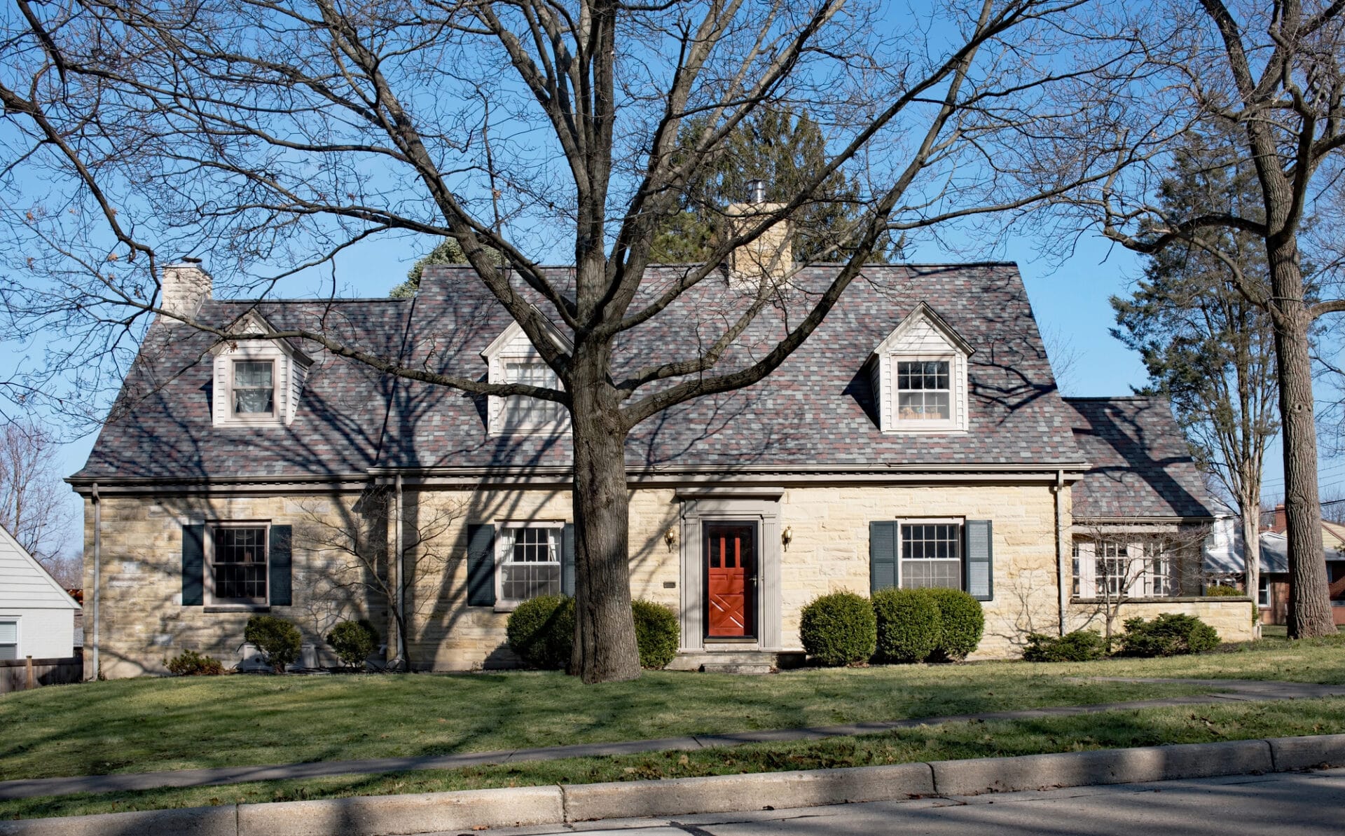 A stone house with a gray shingle roof, red front door, dormer windows, and trimmed bushes stands behind a large leafless tree on a sunny day. The lawn is green and the sky is clear and blue. - RoofZen, Chester County PA Roofing, Siding and More