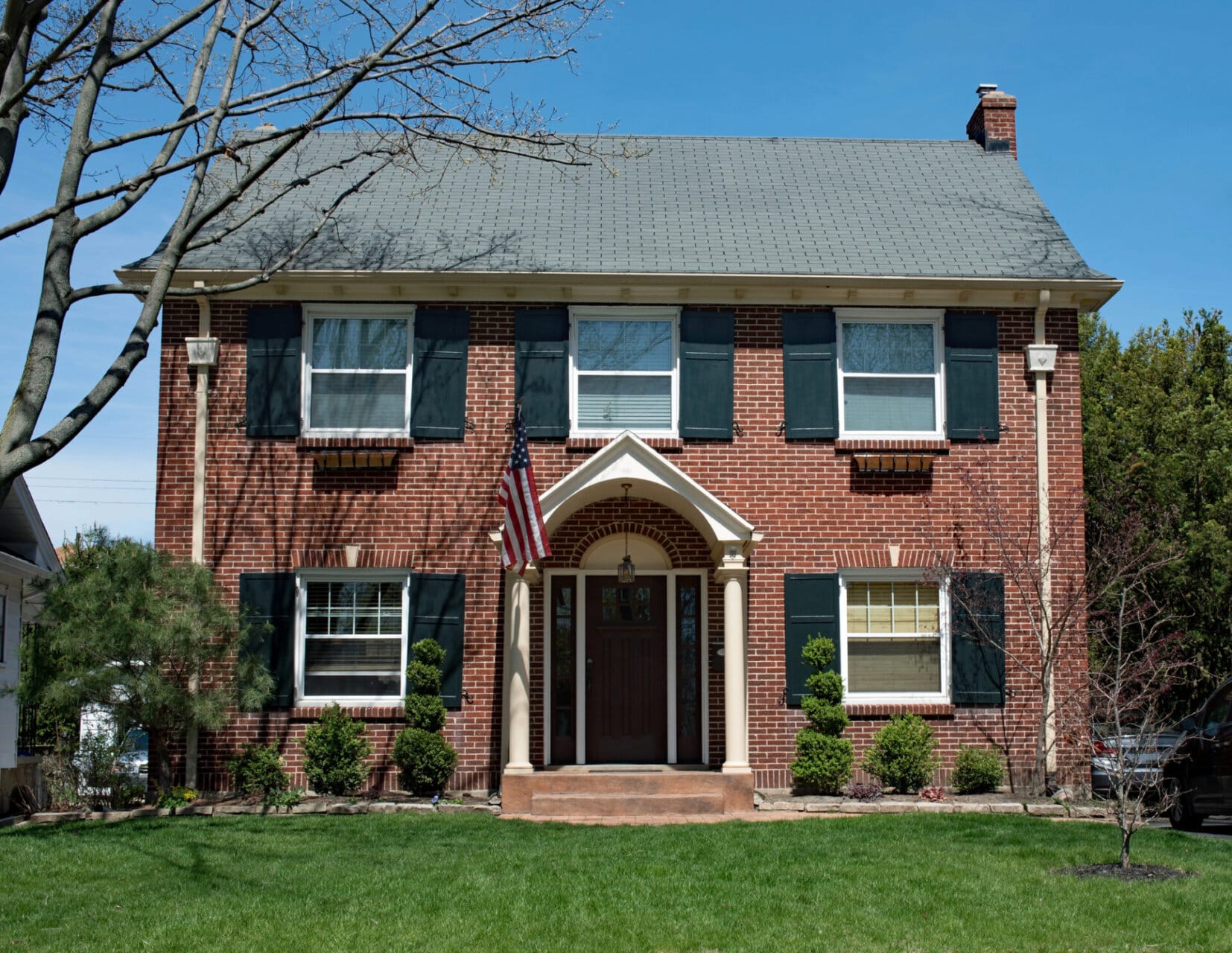 A two-story brick house with black shutters, white trim, and a centered front door under an arched porch. An American flag hangs beside the doorway, and the front yard has green grass and neatly trimmed bushes. - RoofZen, Chester County PA Roofing, Siding and More