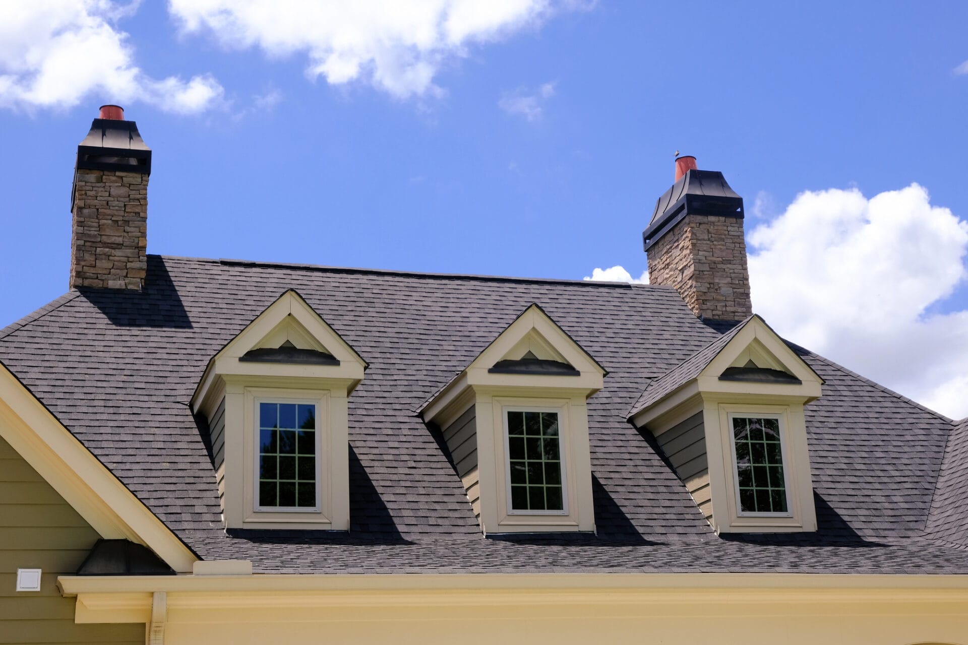 A close-up of a house roof with dark shingles, three dormer windows, and two stone chimneys against a blue sky with scattered white clouds. - RoofZen, Chester County PA Roofing, Siding and More