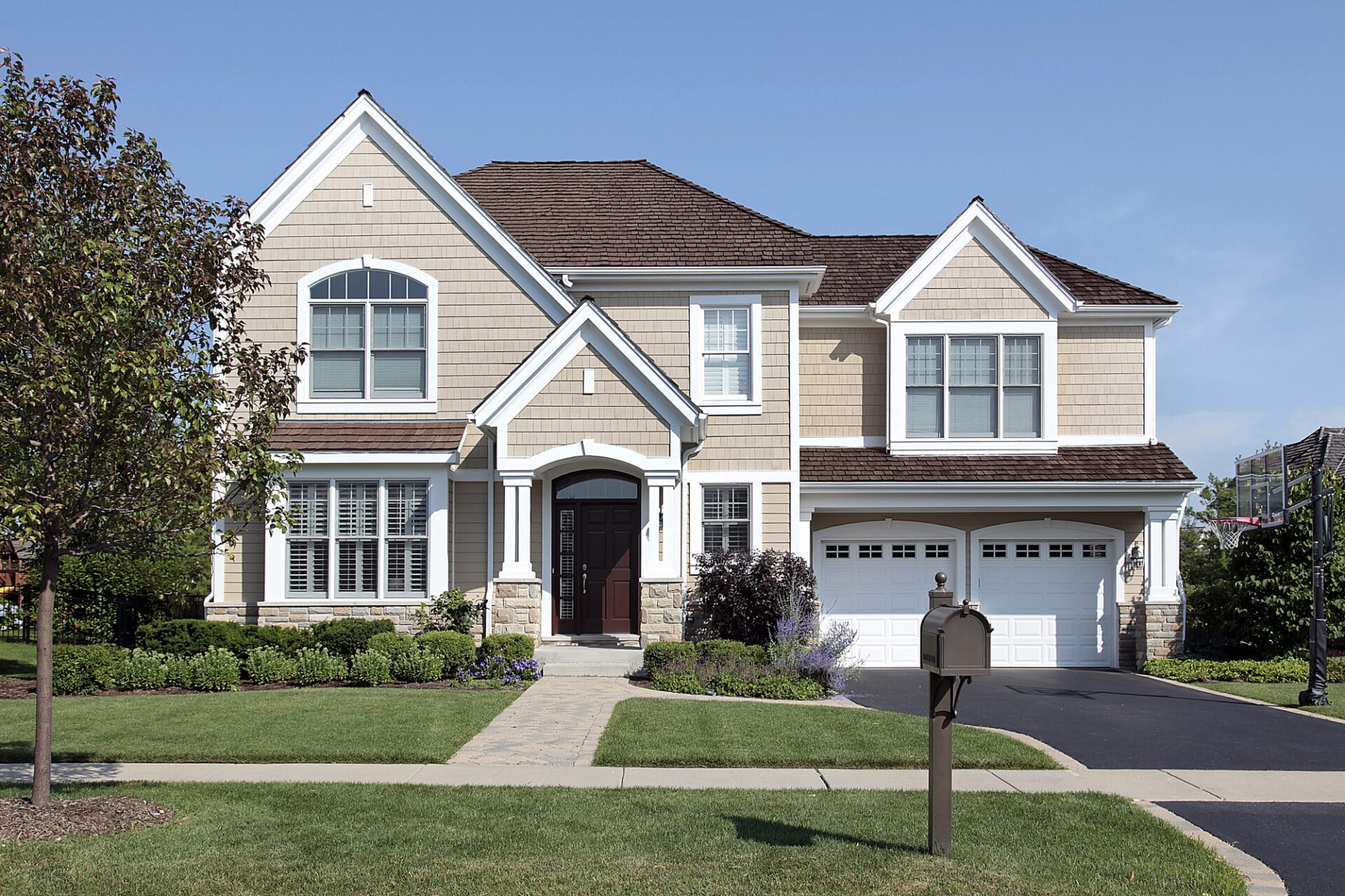 Two-story suburban house with beige siding, white trim, and a dark brown front door. There is a double garage, neatly trimmed bushes, a mailbox, and a tree in the landscaped front yard. The sky is clear and blue. - RoofZen, Chester County PA Roofing, Siding and More