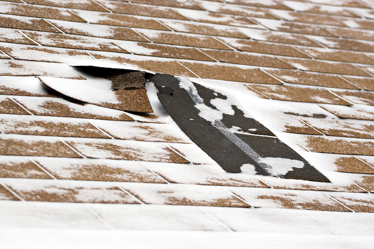 A close-up of a roof with brown shingles covered in patches of snow and a large section of damaged and missing shingles, exposing the black underlayment beneath. - RoofZen, Chester County PA Roofing, Siding and More