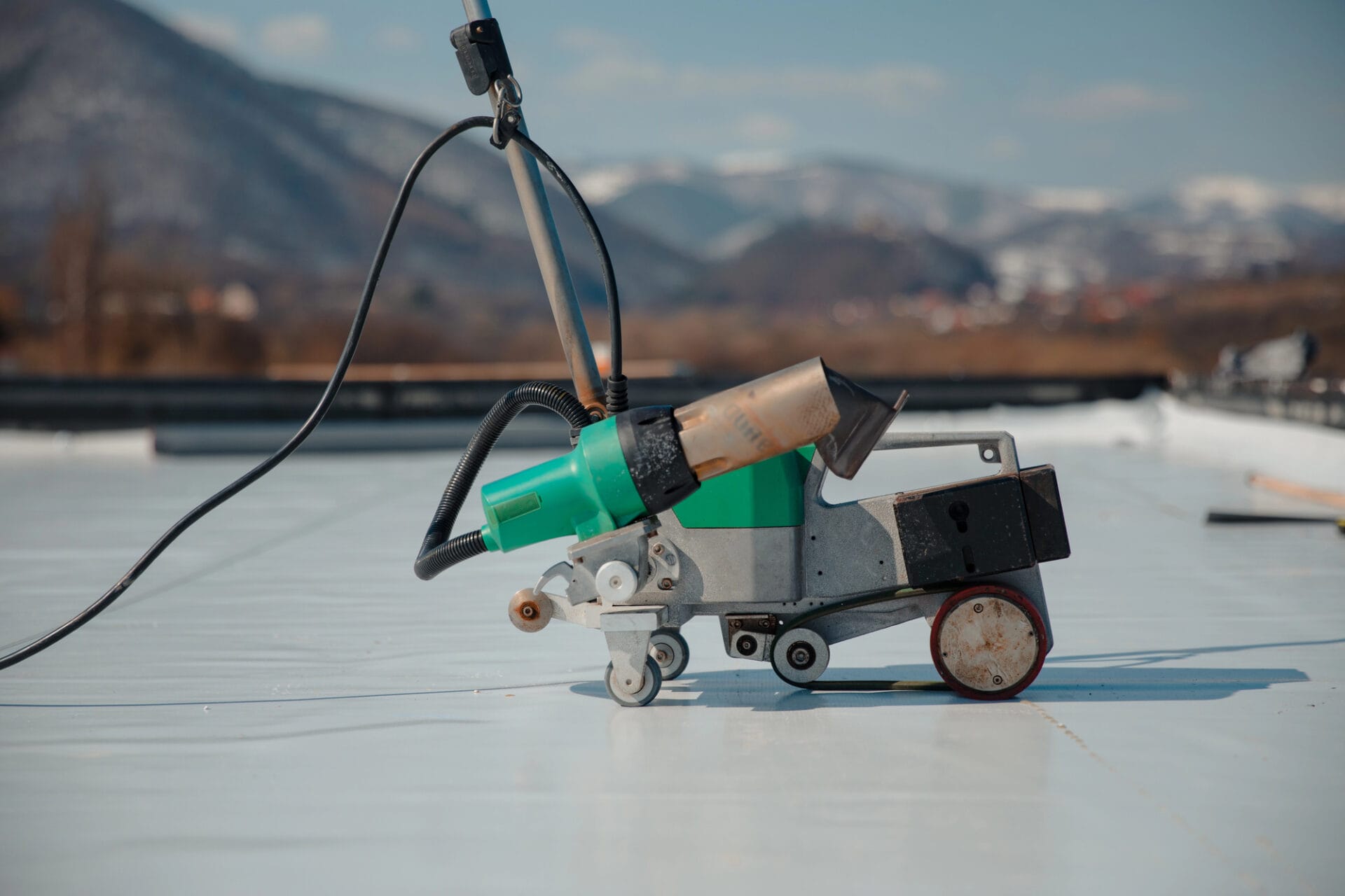 A close-up of a green and black hot air welding machine on a flat white rooftop, with mountains and a blurred landscape in the background. - RoofZen, Chester County PA Roofing, Siding and More