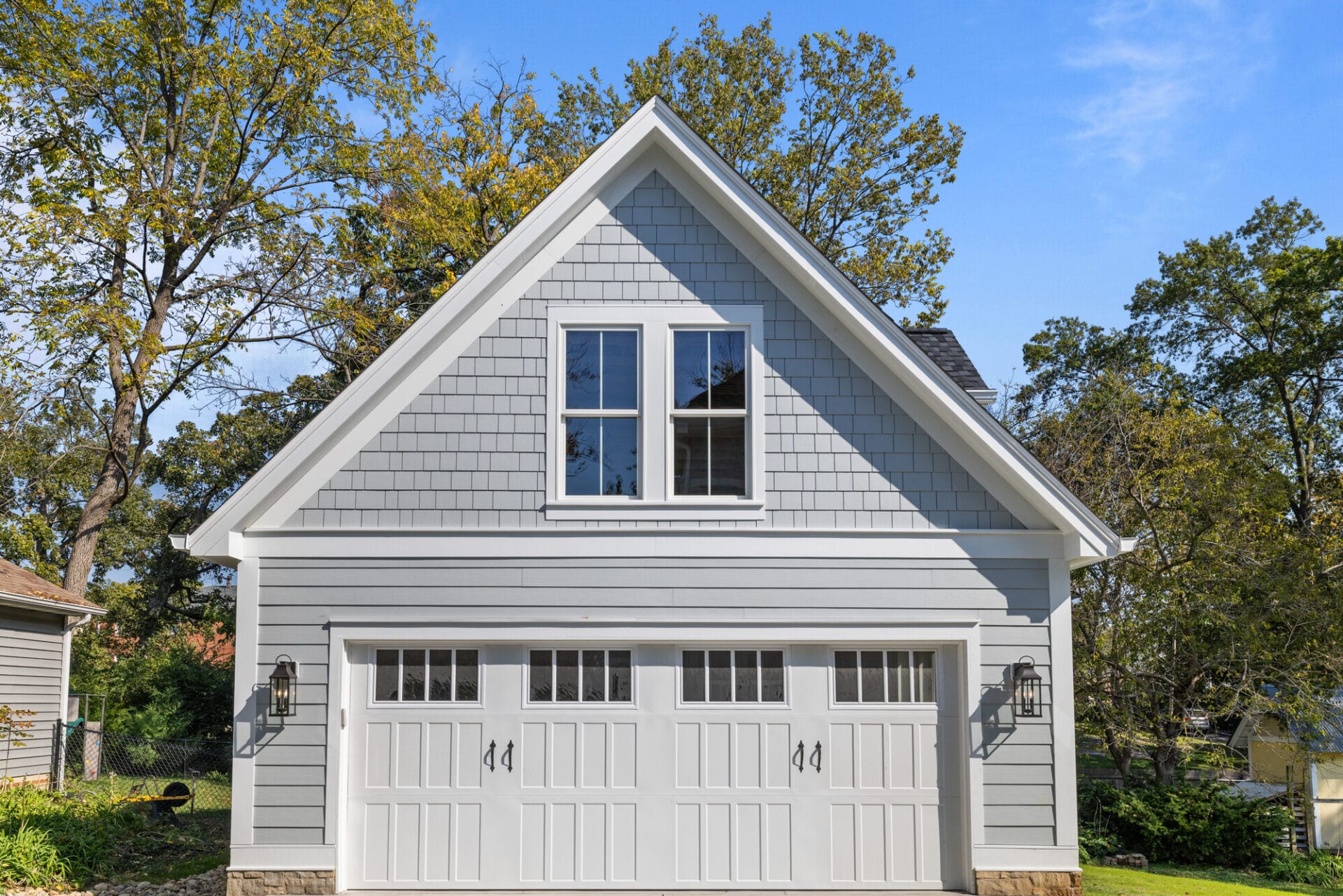 A detached two-car garage with light gray siding, white trim, and an upper window, set among leafy trees on a clear day. - RoofZen, Chester County PA Roofing, Siding and More