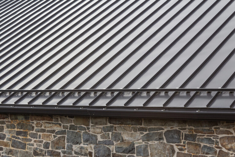 Close-up view of a modern metal roof with standing seam panels above a wall made of irregularly shaped stone bricks. The roof features clean, parallel lines and a dark finish. - RoofZen, Chester County PA Roofing, Siding and More