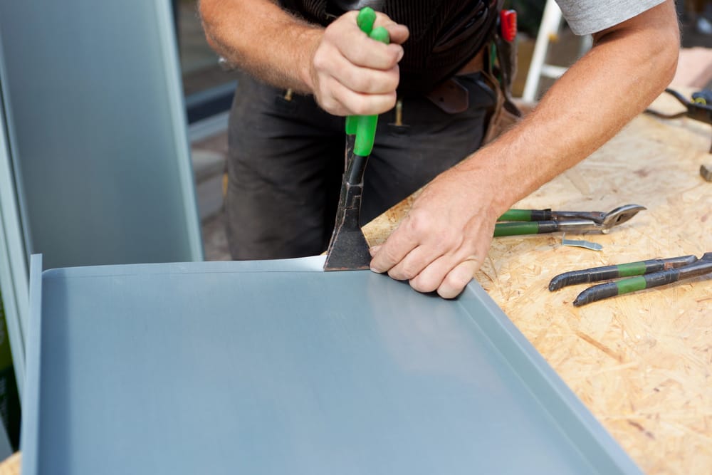 A person uses a green-handled tool to bend or shape a sheet of metal on a wooden workbench. Other hand tools are visible nearby on the table. - RoofZen, Chester County PA Roofing, Siding and More